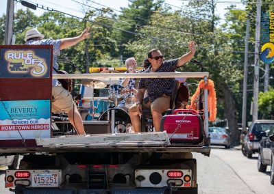 Rule of 3 band members waving from the back of the truck with a Beverly Homecoming Honors Our Hometown Heroes sign